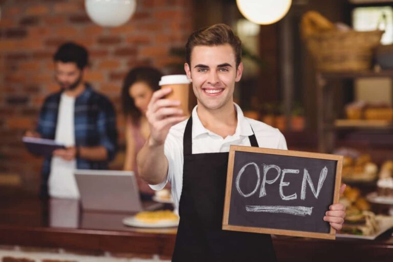 Soar smiling barista holding coffee and open sign laptop users in background