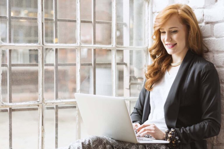 Soar woman sitting against brick wall next to window typing on laptop on her lap